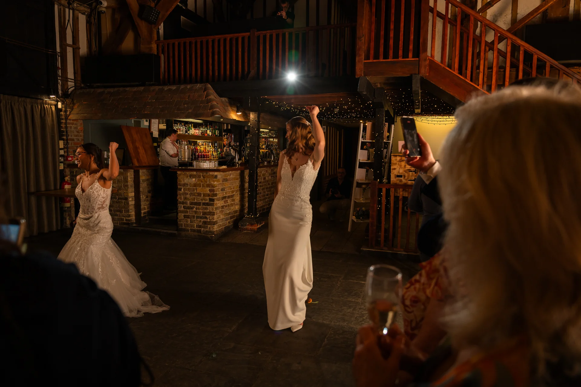 Two brides in stunning wedding dresses dance joyfully on the dance floor of a rustic barn reception venue at night, surrounded by cheering guests.