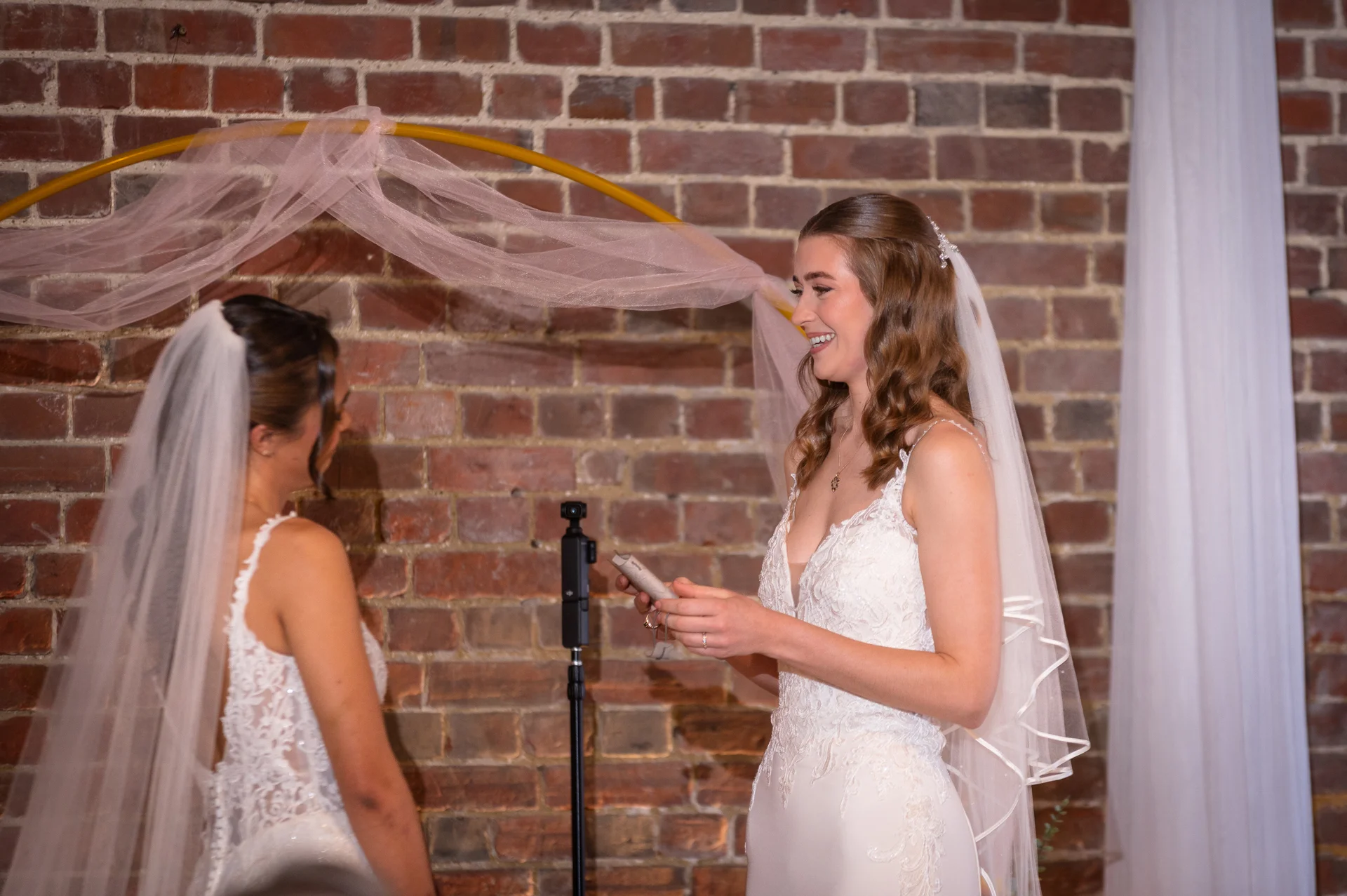 Two smiling brides stand opposite each other during their outdoor wedding ceremony in front of an exposed brick wall and a decorative pink-draped arch.