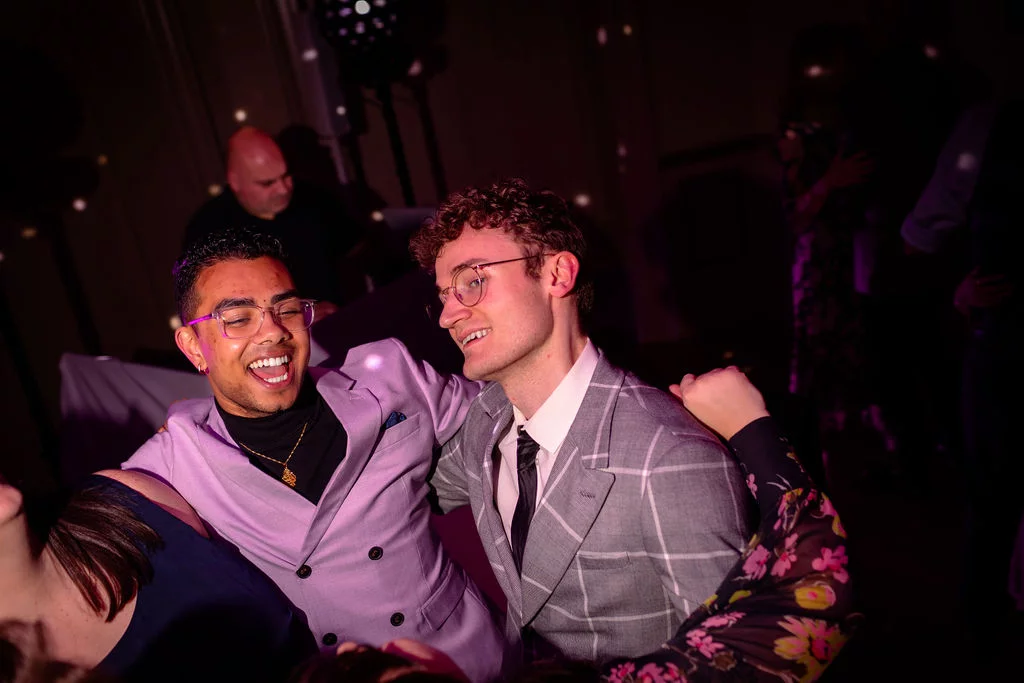 Two smiling men embrace and laugh together on a crowded dance floor bathed in purple light during a wedding reception.