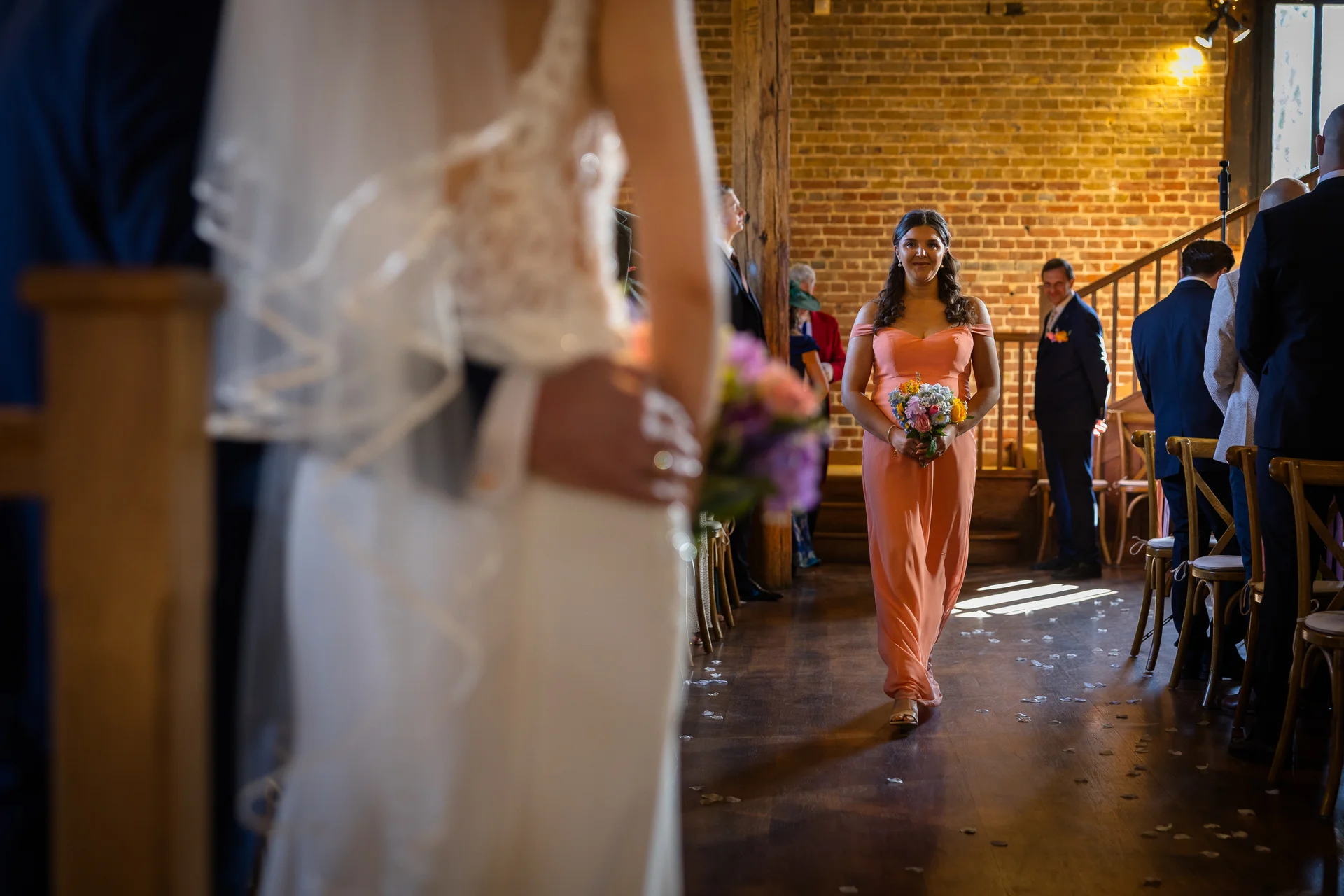 A bridesmaid in a coral dress walks down the aisle inside a rustic brick barn, viewed from behind the bride standing at the front.