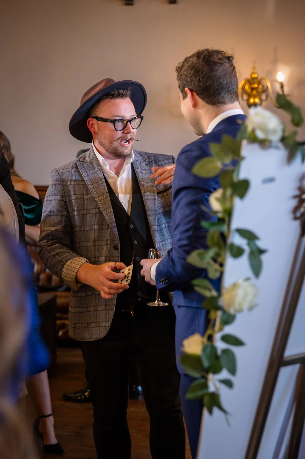 A close up magician performs a card trick for a wedding guest wearing a checked suit and fedora during the drinks reception.