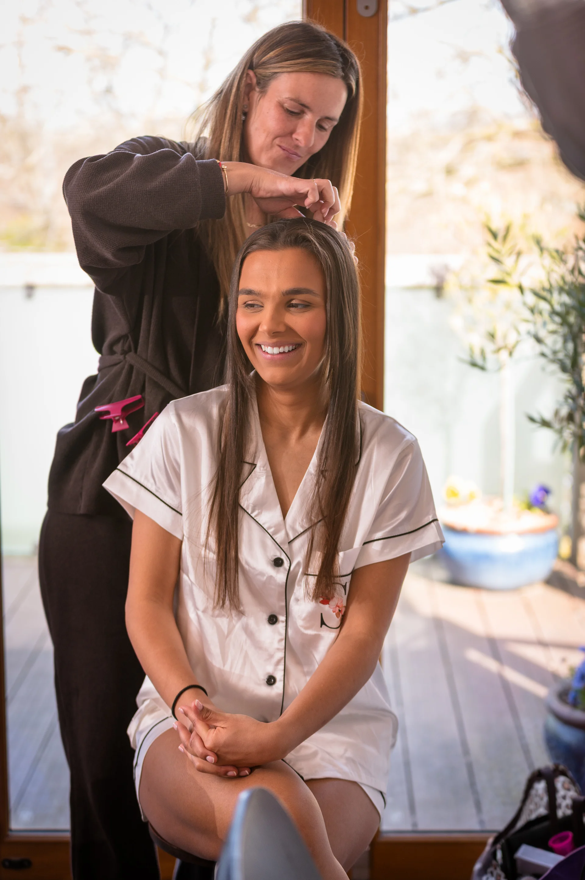 A smiling bride wearing white satin pyjamas has her long dark hair styled by a hairdresser during morning bridal preparations near a bright window.
