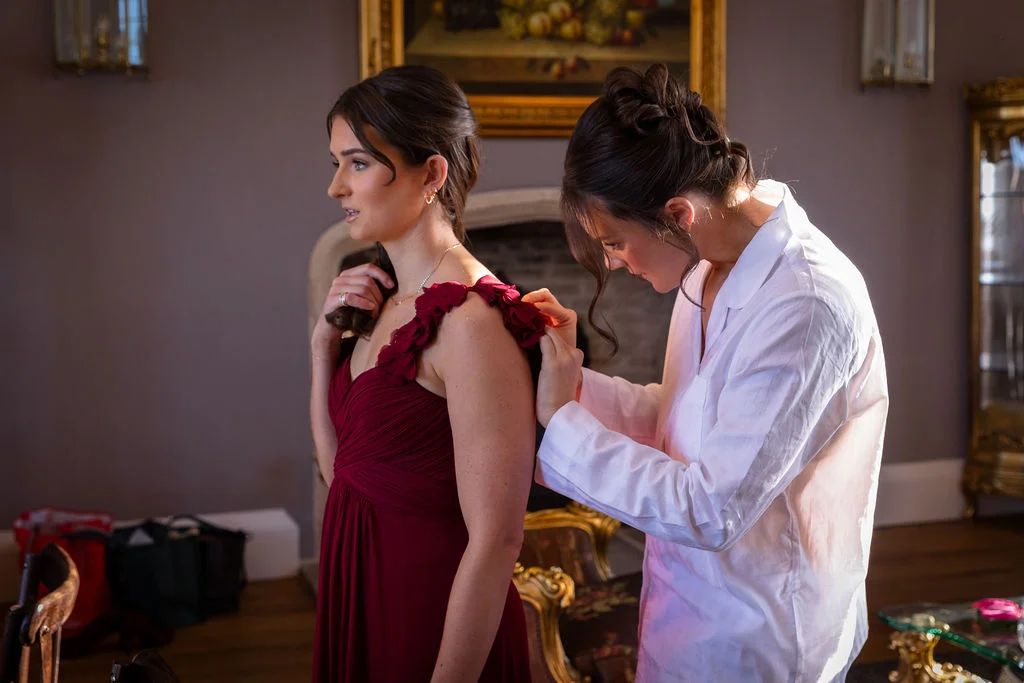 A bridesmaid in a matching white shirt assists another bridesmaid with adjusting her pleated burgundy dress during morning preparations.