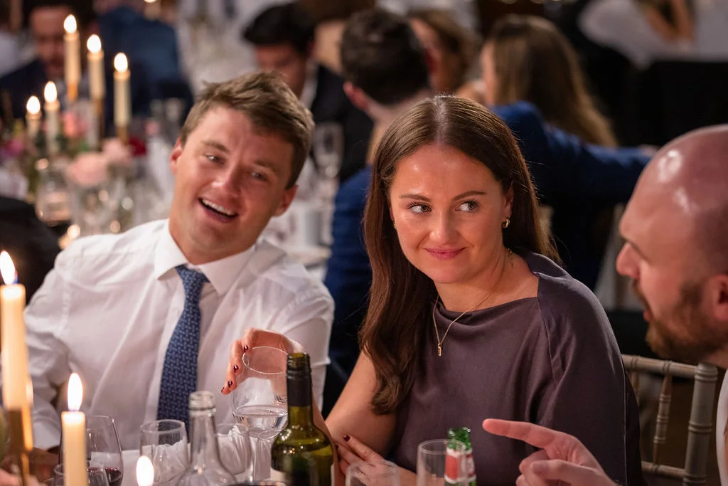 Candid photograph of wedding guests smiling and mingling around a candlelit dining table during the wedding breakfast.