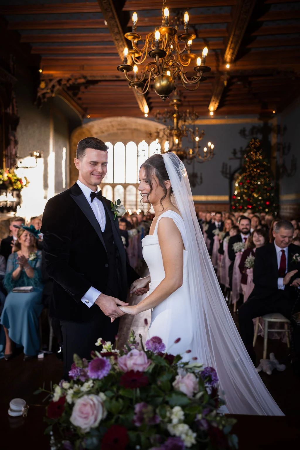 A newlywed couple holds hands at the altar during their elegant ceremony inside a historic hall lit by grand chandeliers.