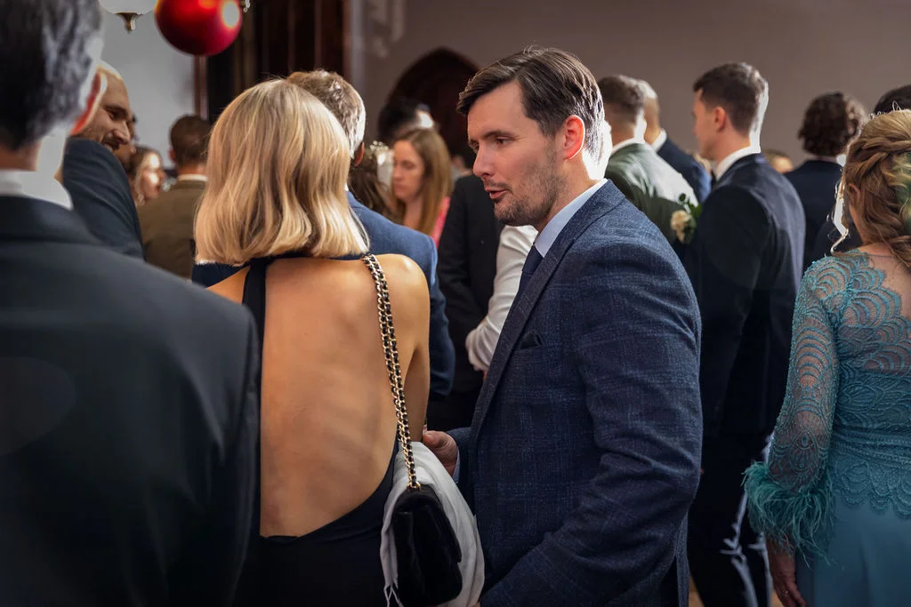 Wedding guests mingle during a lively drinks reception, with a man in a navy suit conversing with a guest in an elegant black dress.