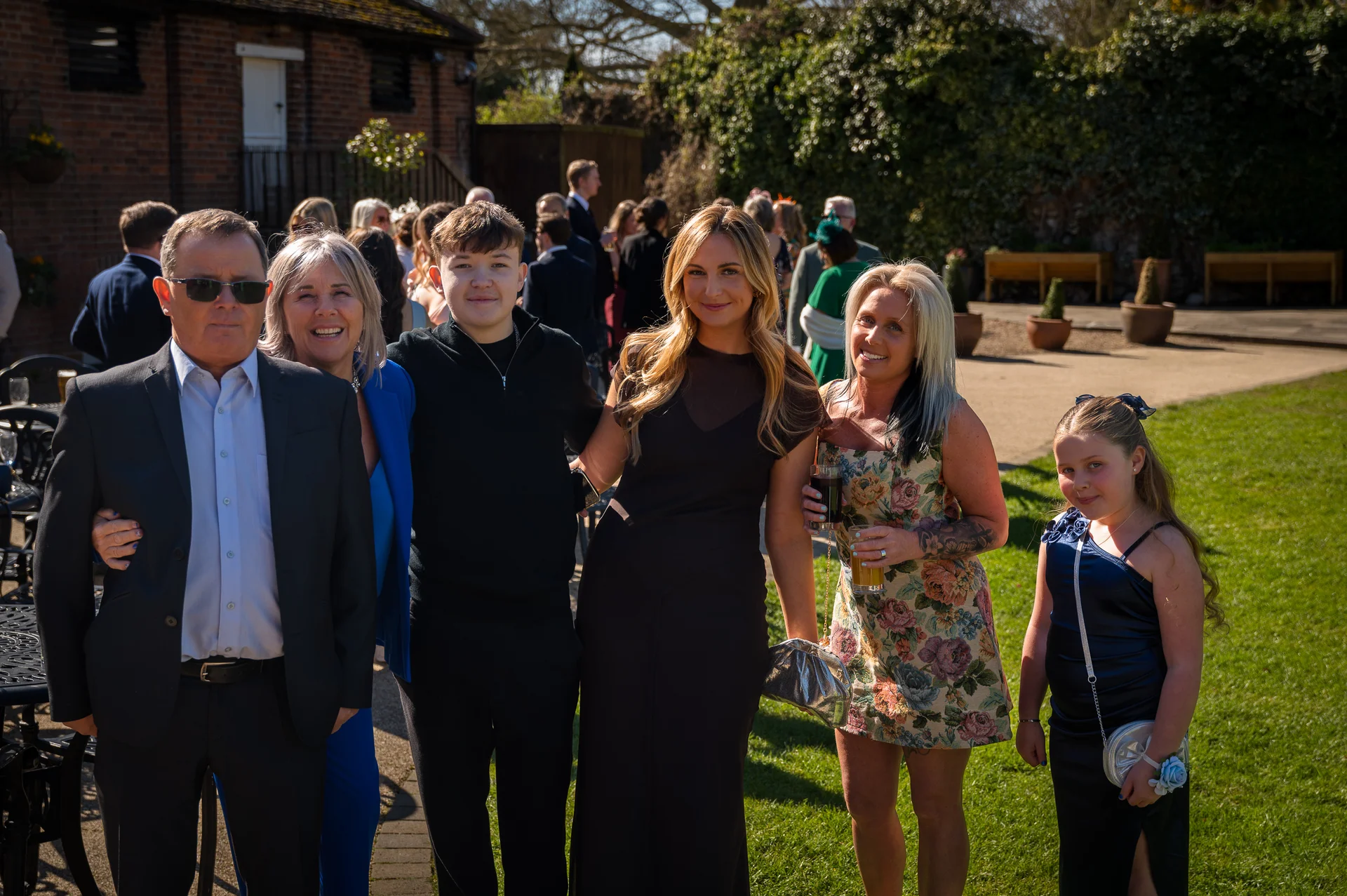 A group of wedding guests pose together outdoors in a sunny courtyard with a brick and greenery backdrop.
