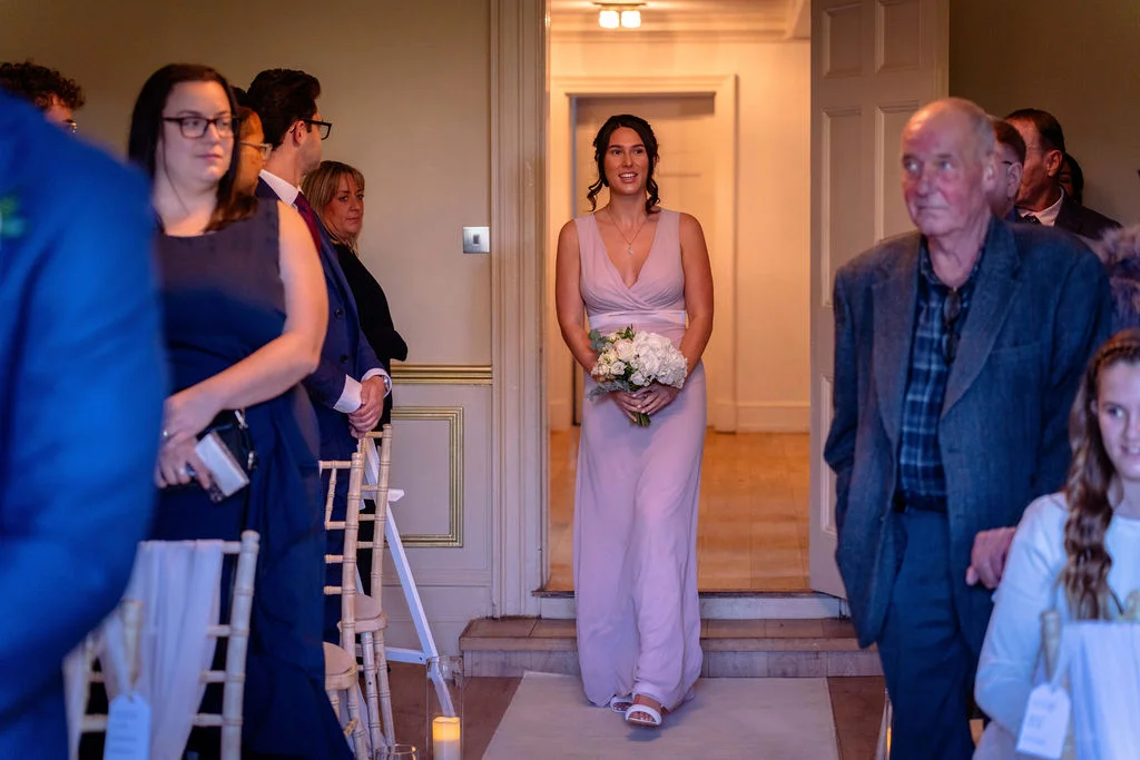 A smiling bridesmaid in a dusty pink dress holding a white bouquet walks down the aisle during a wedding processional.