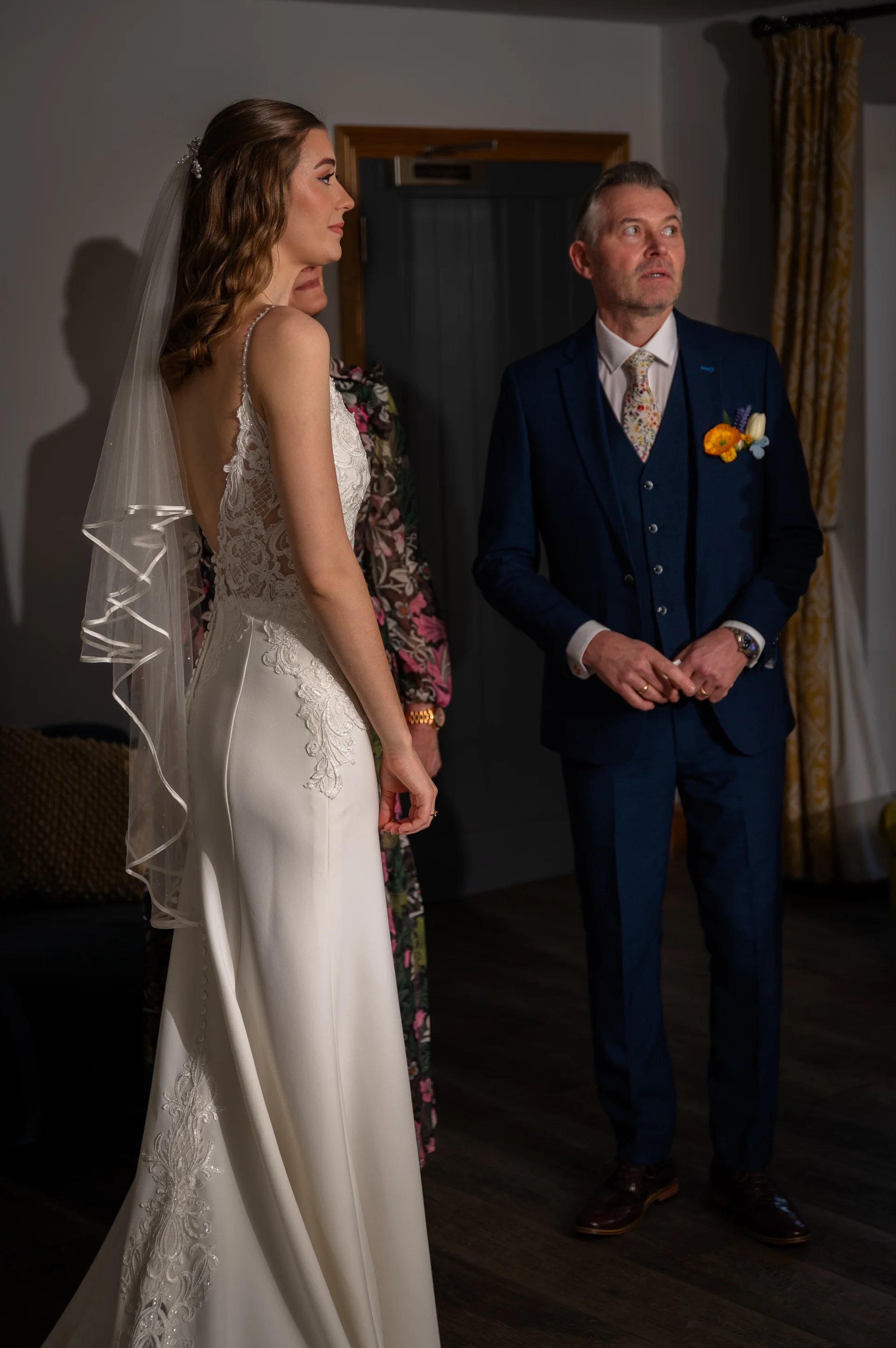 A stunning bride in a form-fitting crepe wedding gown with a lace-detailed open back and veil stands beside her father in a smart navy three-piece suit.
