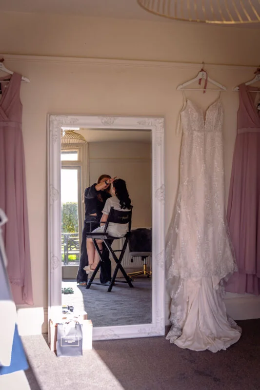 A bride sits in a director chair getting her makeup applied while reflected in a large ornate mirror, flanked by her hanging wedding dress and pink bridesmaid dresses.