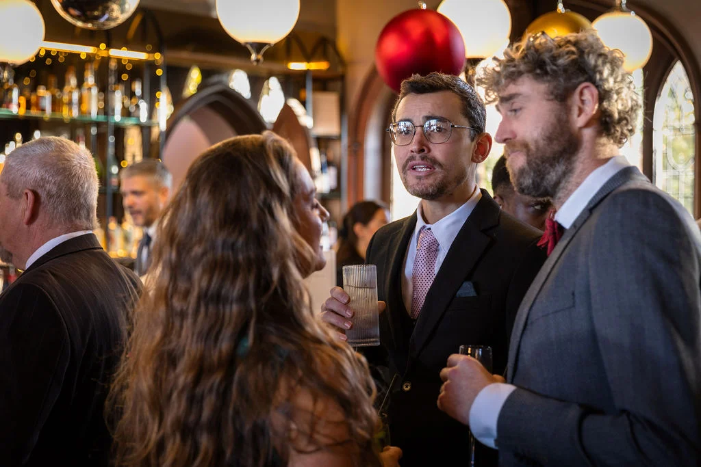 Wedding guests mingling and enjoying drinks inside a lively pub reception, with two men in suits chatting animatedly.