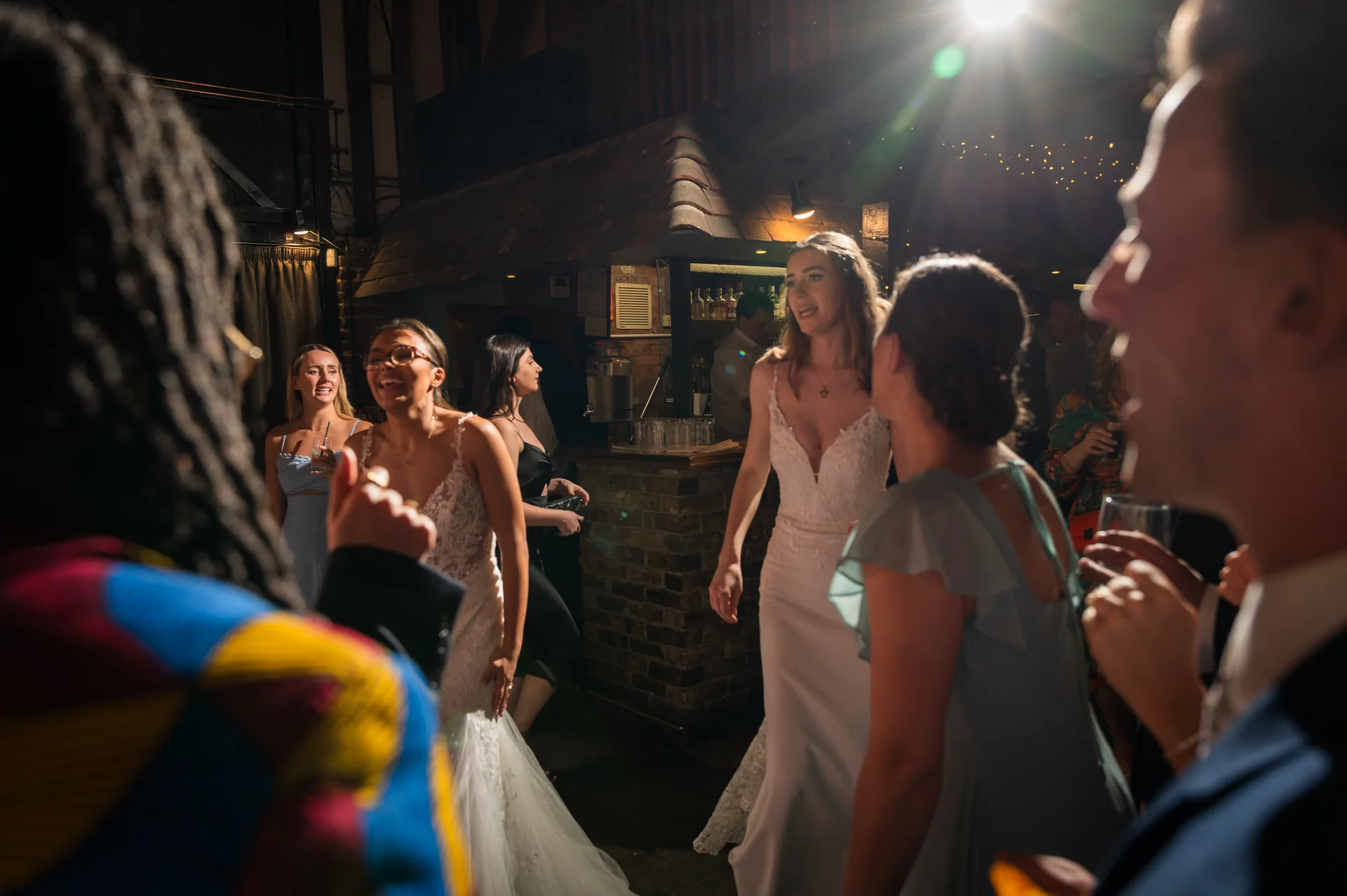 A candid evening shot of the bride laughing and dancing with guests on a crowded dance floor in a pub venue illuminated by dramatic spotlighting.