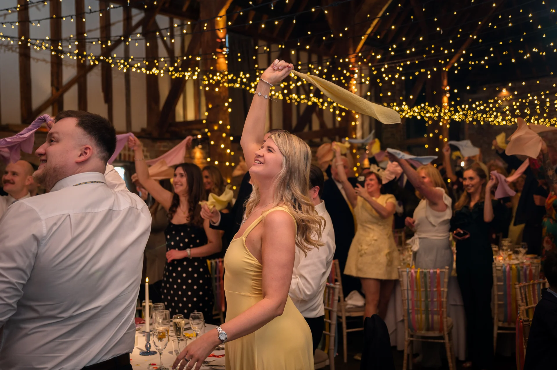 Wedding guests enthusiastically wave napkins in the air during a lively barn reception illuminated by a warm fairy light canopy.