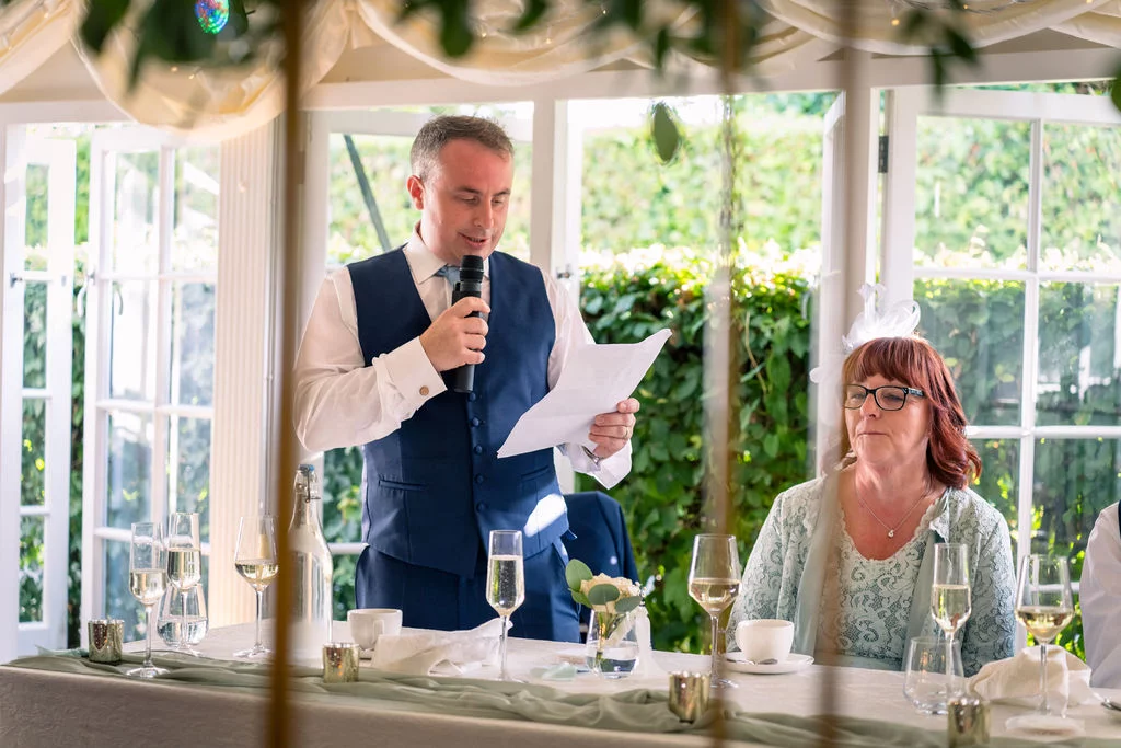 A groom in a blue waistcoat delivers his wedding speech holding notes and a microphone, seated next to the mother of the bride at a beautifully styled banquet table in a sunlit orangery.