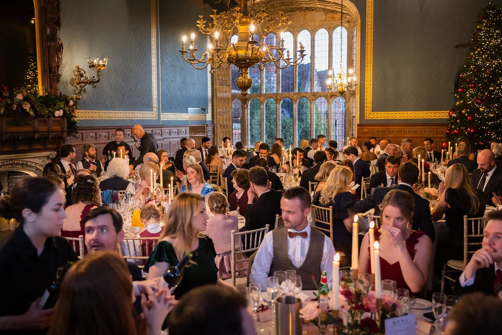 Wedding guests enjoy a festive wedding breakfast inside a grand stately home banquet hall adorned with a lit Christmas tree and candlelit tables.