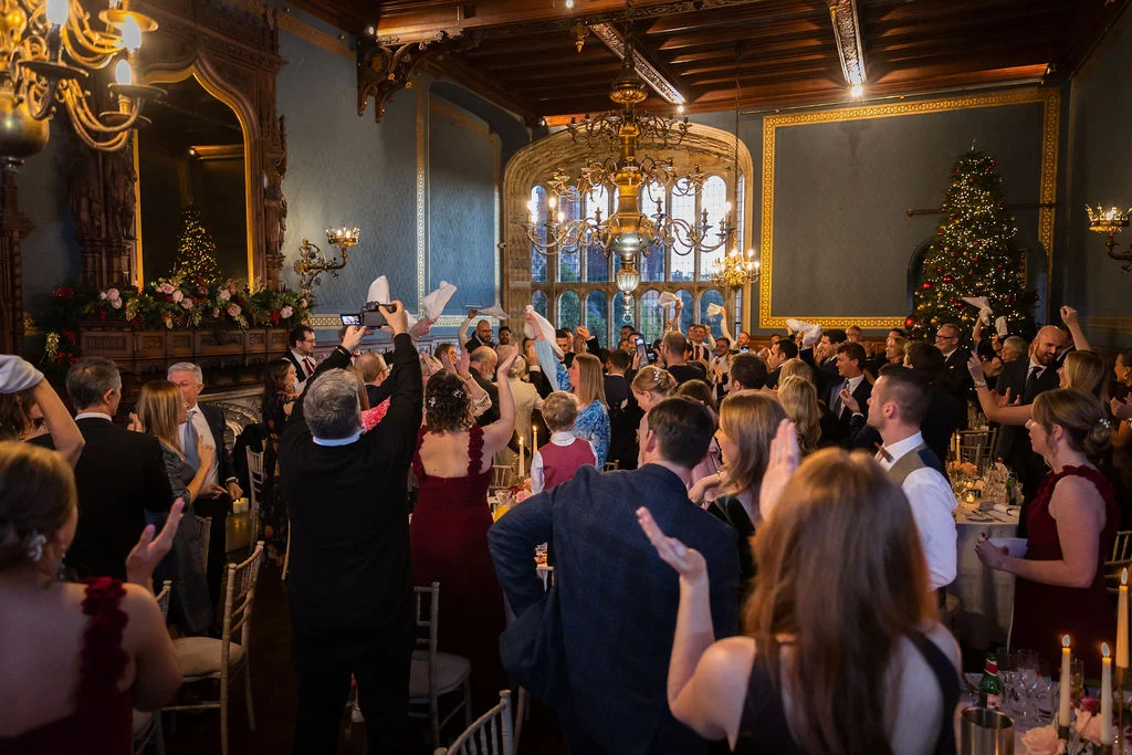 Wedding guests enthusiastically wave napkins in the air during the wedding breakfast inside a grand, wood-panelled ballroom decorated for Christmas.