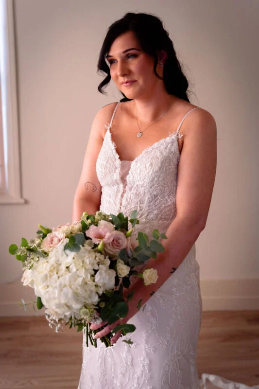 A solo bride with dark hair in a half-up style wears a lace spaghetti-strap gown and holds a lush hand-tied bouquet of white hydrangeas, roses, and eucalyptus.