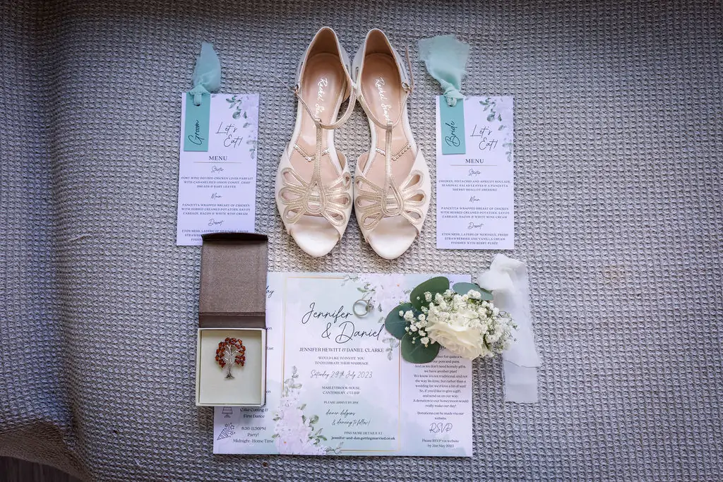 An overhead flat lay showcases a bridal wedding invitation suite featuring floral motifs, matching menus with sage ribbons, metallic t-bar heels, a ring, and a boutonniere with a white rose.