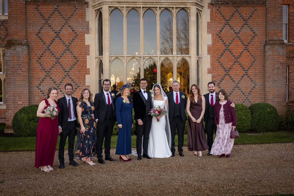 A bridal party and family members pose together outside a grand brick country house featuring large arched windows and well-manicured topiary.
