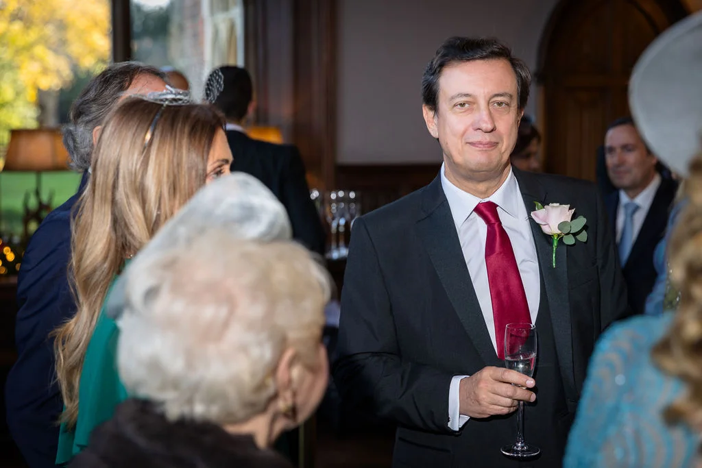 A mature gentleman in a black suit and red tie holds a glass of champagne while mingling with guests at a wedding drinks reception.