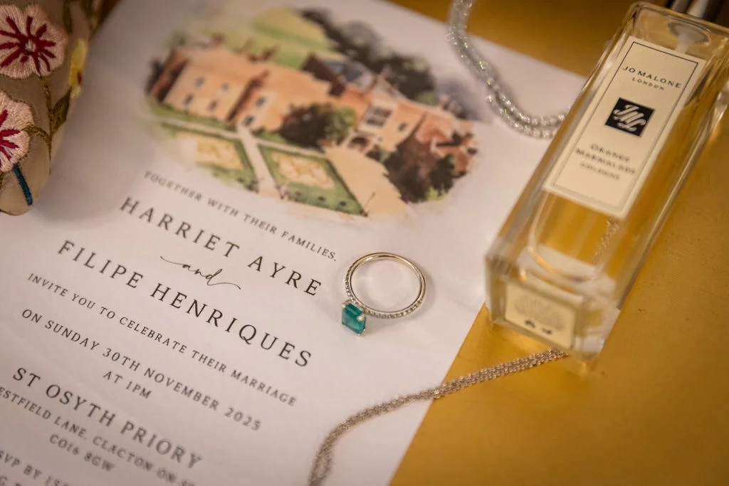 An elegant flat lay of a wedding invitation featuring a venue illustration, accompanied by a sparkling ring with a green gemstone, a perfume bottle, and delicate silver jewelry.