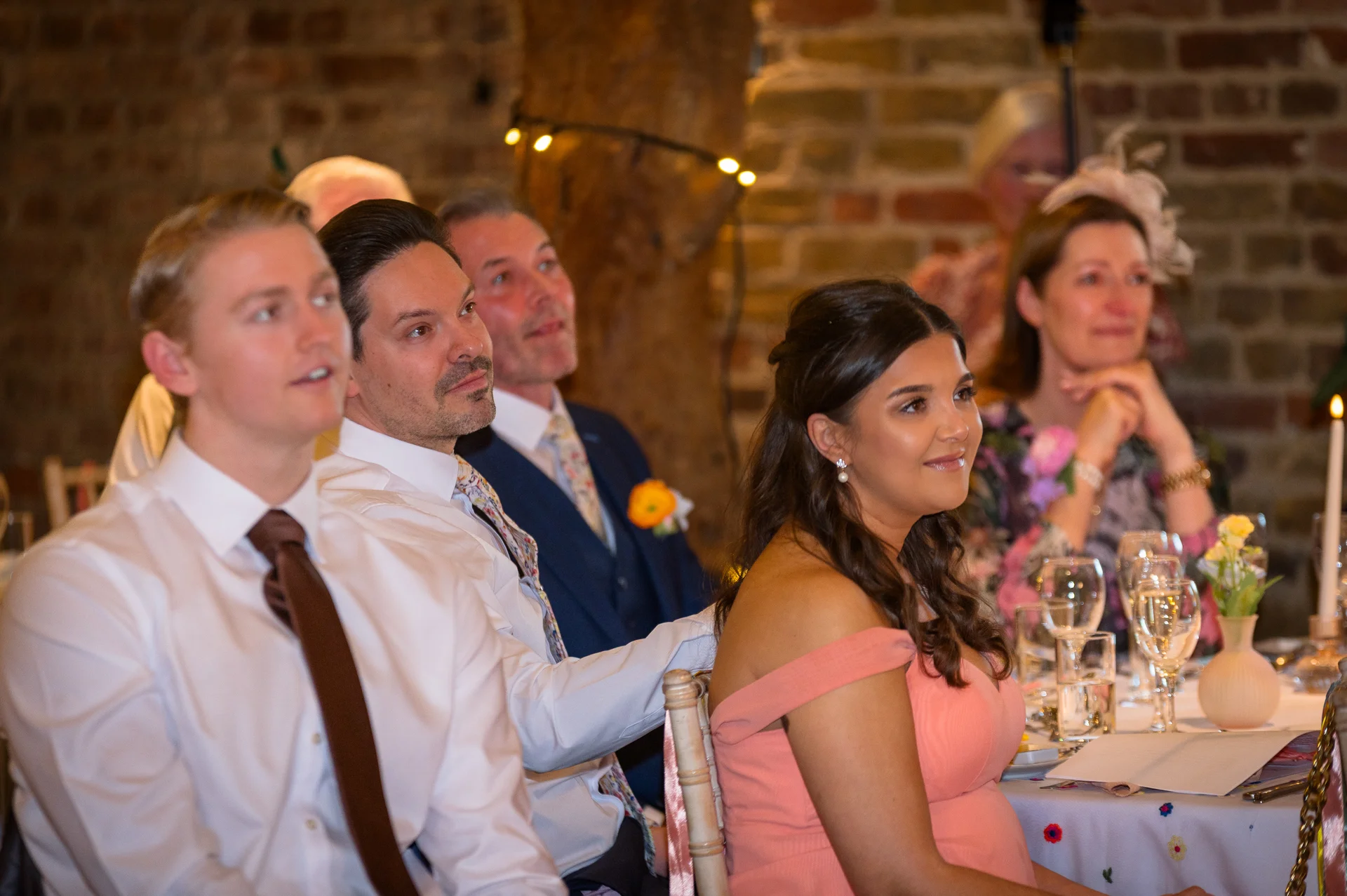 Wedding guests smiling and listening attentively during speeches at a rustic barn reception decorated with candles.