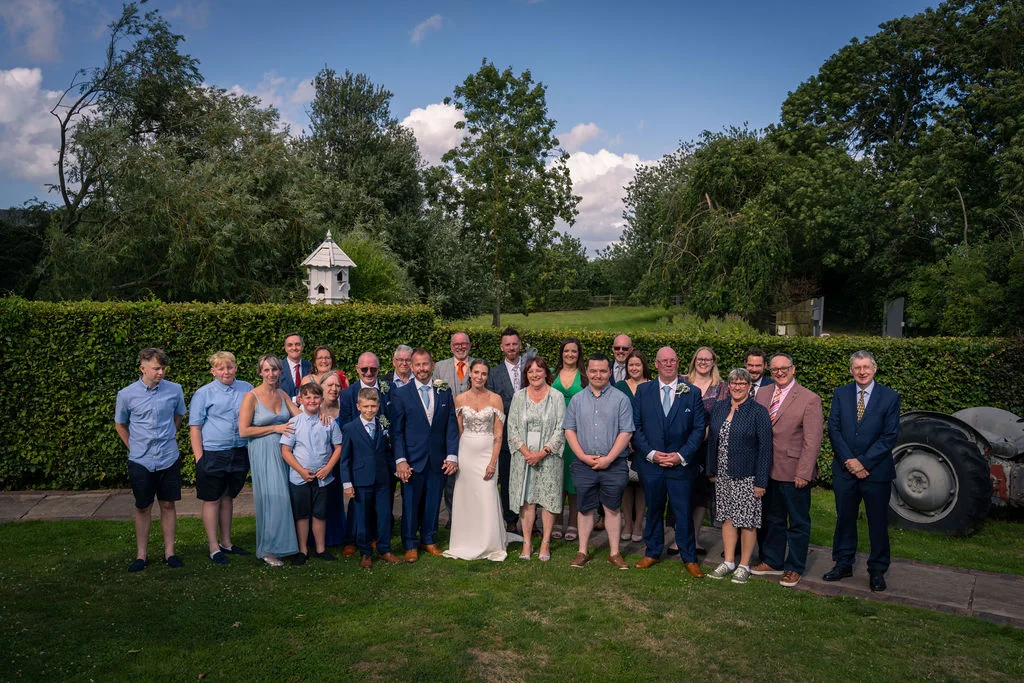 A large wedding party and family group pose together outdoors on a manicured lawn against a backdrop of lush green hedges and tall trees under a bright blue sky.