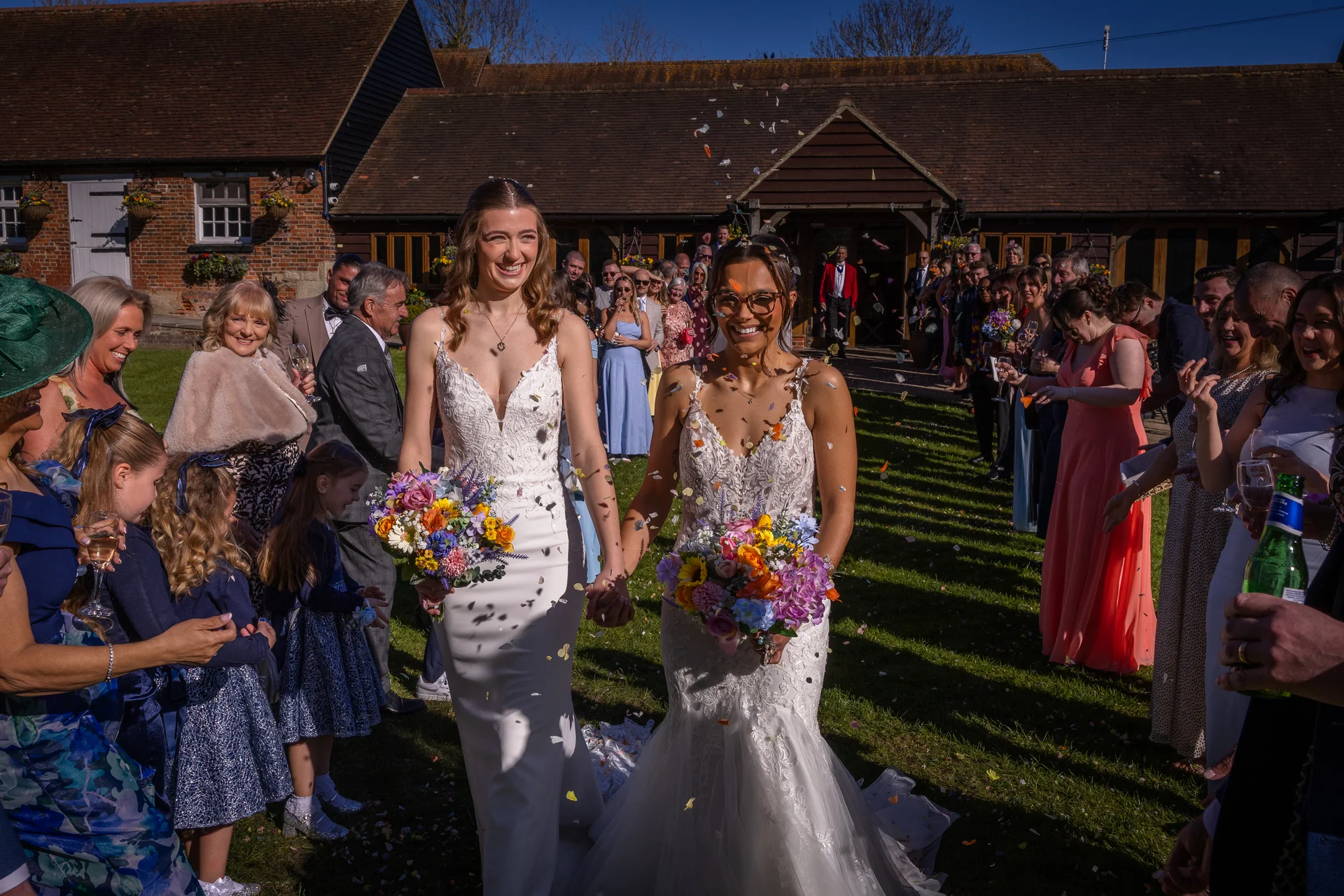Two smiling brides holding colorful bouquets walk hand-in-hand through a cheering crowd of wedding guests tossing confetti outside a rustic stone barn.
