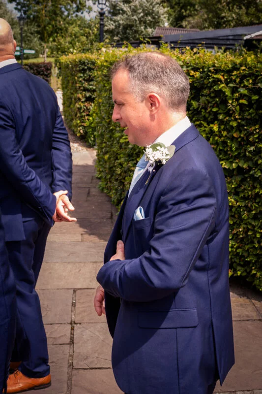 A groom smiling in profile while wearing a navy blue suit, light blue tie, and a delicate floral buttonhole as he stands near a green garden hedge.