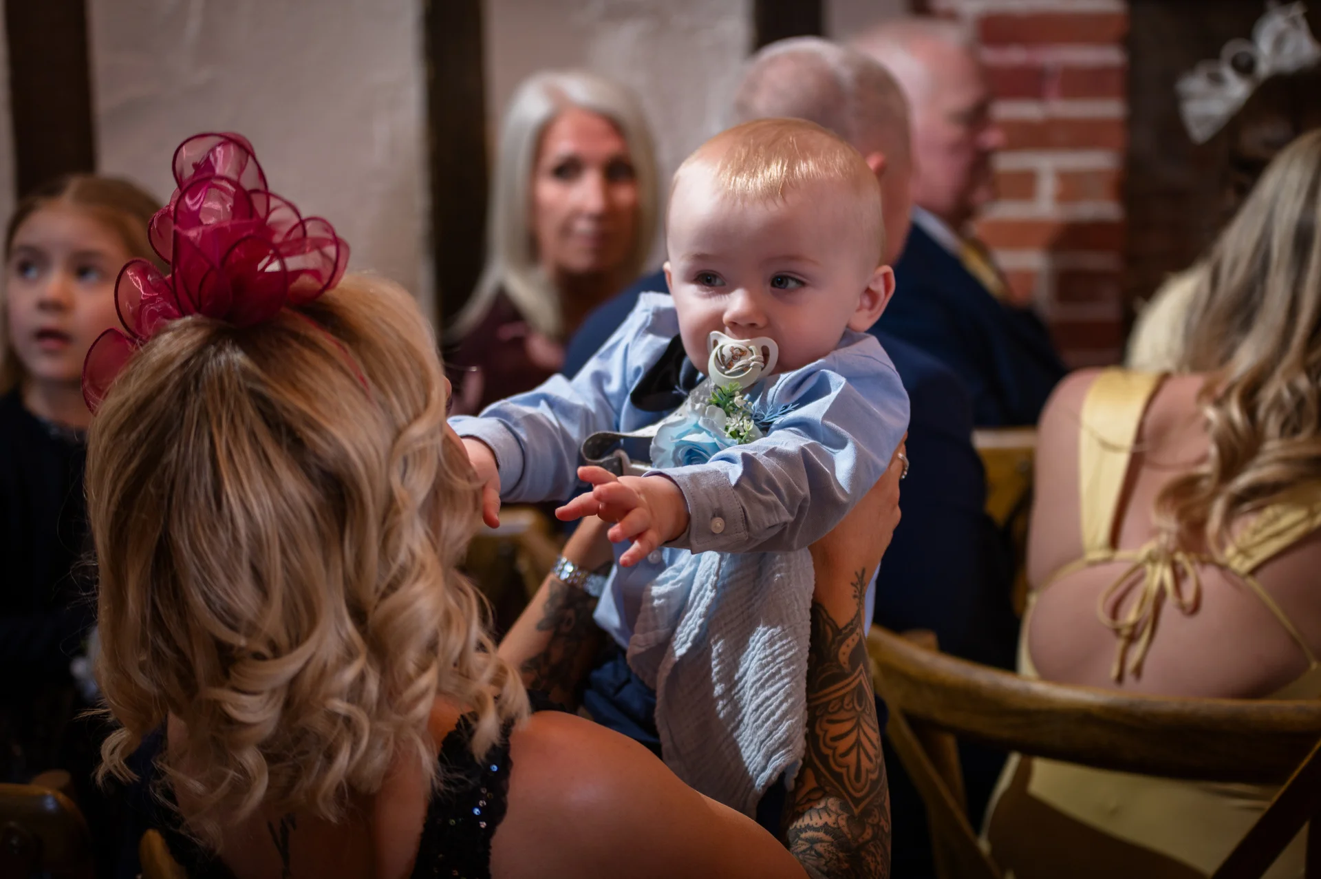 A tattooed guest holds a baby with a dummy at a wedding reception while other guests mingle in the background.