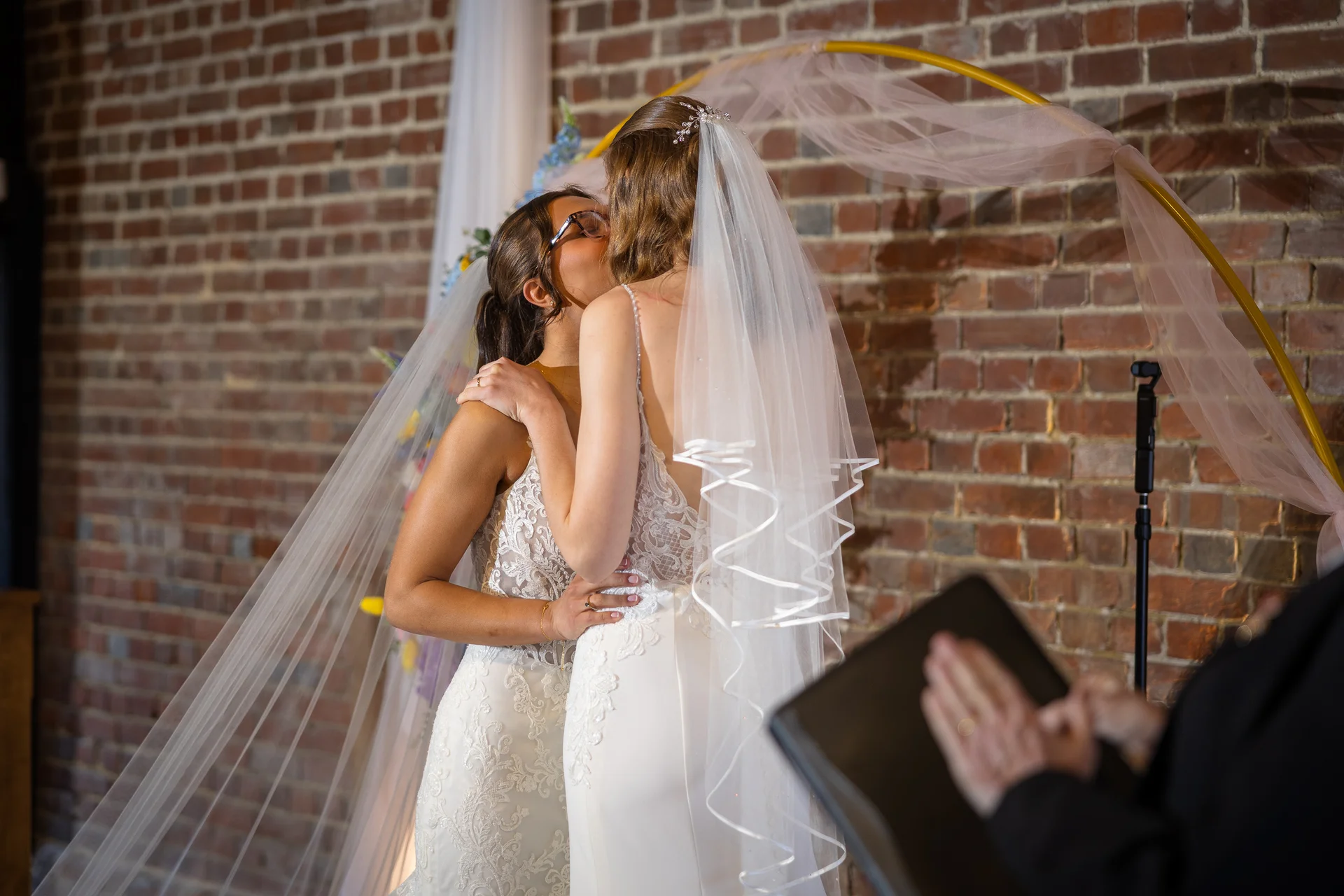 Two brides share their first kiss as a married couple beneath a floral-adorned golden arch in front of an exposed brick wall.