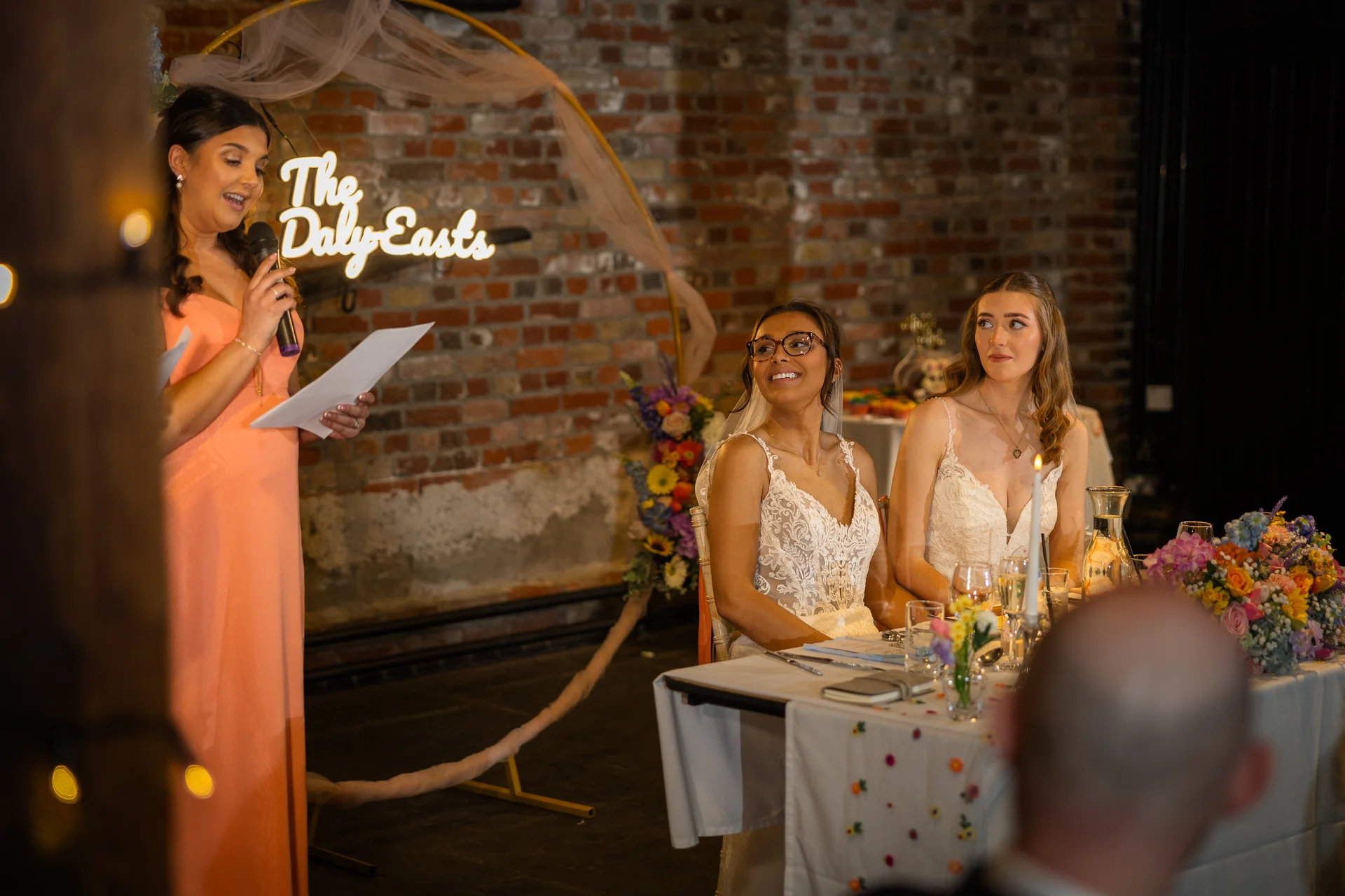 A smiling bride wearing glasses and her wife listen attentively during a heartfelt speech delivered by a bridesmaid at a rustic brick venue.