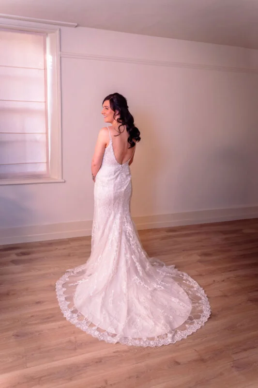A smiling bride stands in a bright room with wooden floors, showcasing the intricate lace detailing and sweep train of her wedding dress from behind.