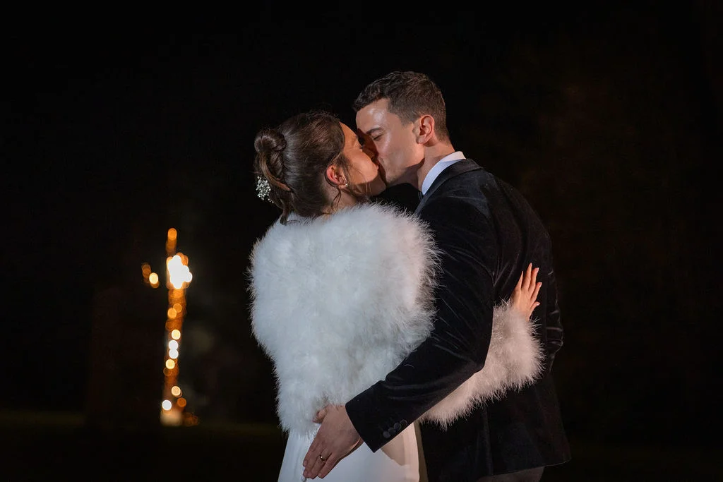 A newlywed couple share a tender kiss at night outdoors, with the bride wearing a plush white faux fur stole and the groom in a black velvet tuxedo.