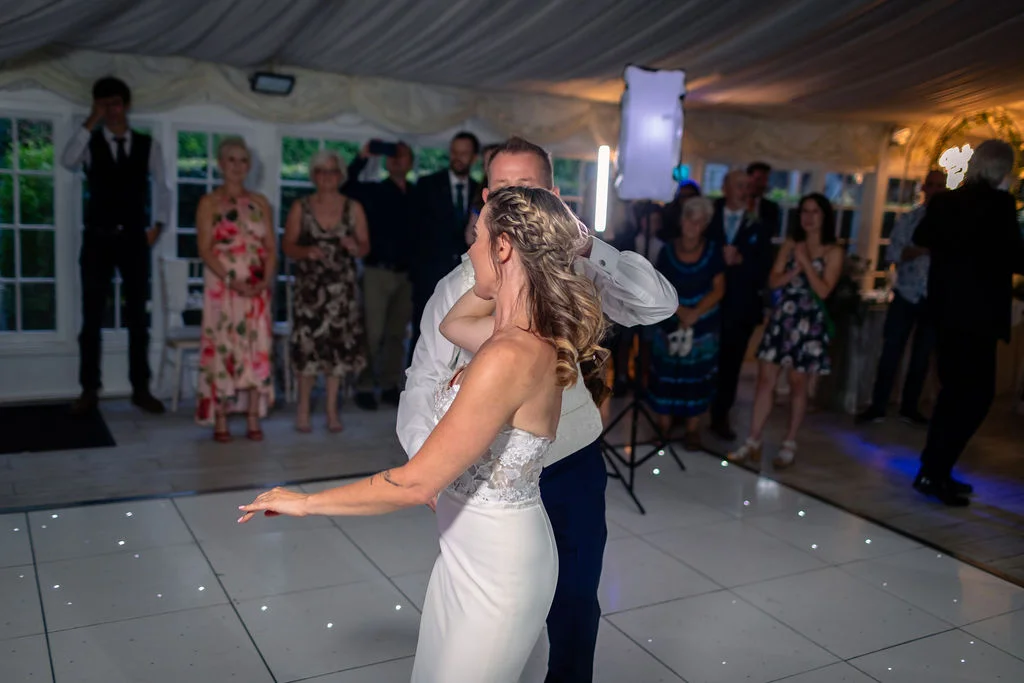 A newlywed couple shares their first dance on a white illuminated dance floor inside a marquee surrounded by watching wedding guests.