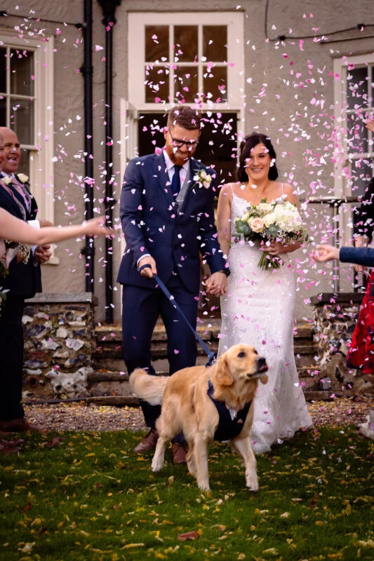 A newlywed couple laugh as pink heart-shaped confetti rains down on them outside a country house, while their golden retriever dog walks excitedly on a lead in the foreground.