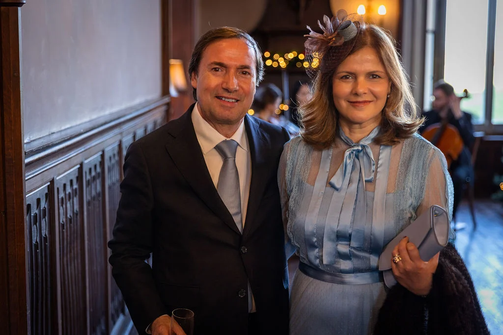 An elegantly dressed older couple smiles together inside a wood-panelled venue during a wedding reception, with the female guest wearing a light blue dress and a matching fascinator.