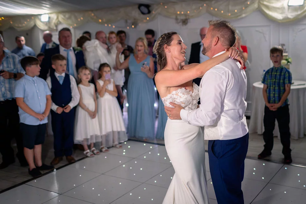 The newlyweds share their first dance on a lighted dance floor while guests and family members watch on with smiles.