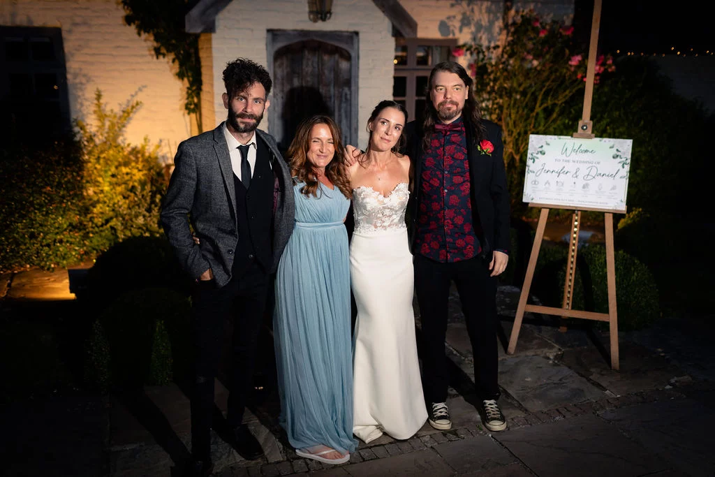 A newlywed couple and two guests pose together outside a lit building at night beside a wooden welcome sign.