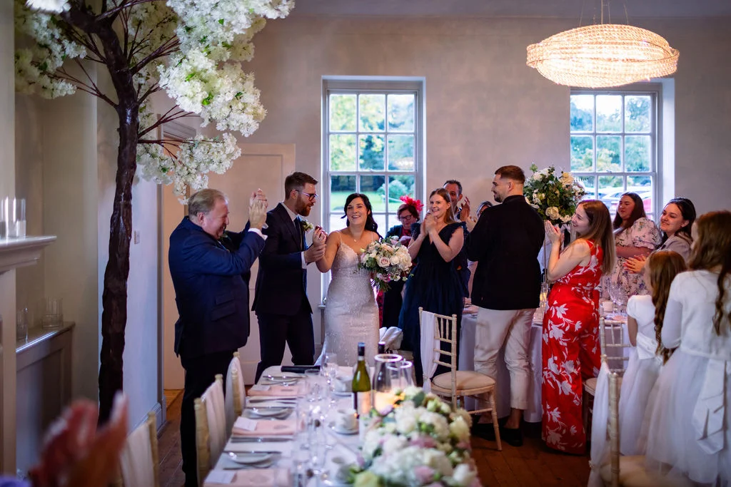 Newlyweds enter a bright reception room with cheering guests, decorative cherry blossom trees, and a beautifully set banquet table in the foreground.