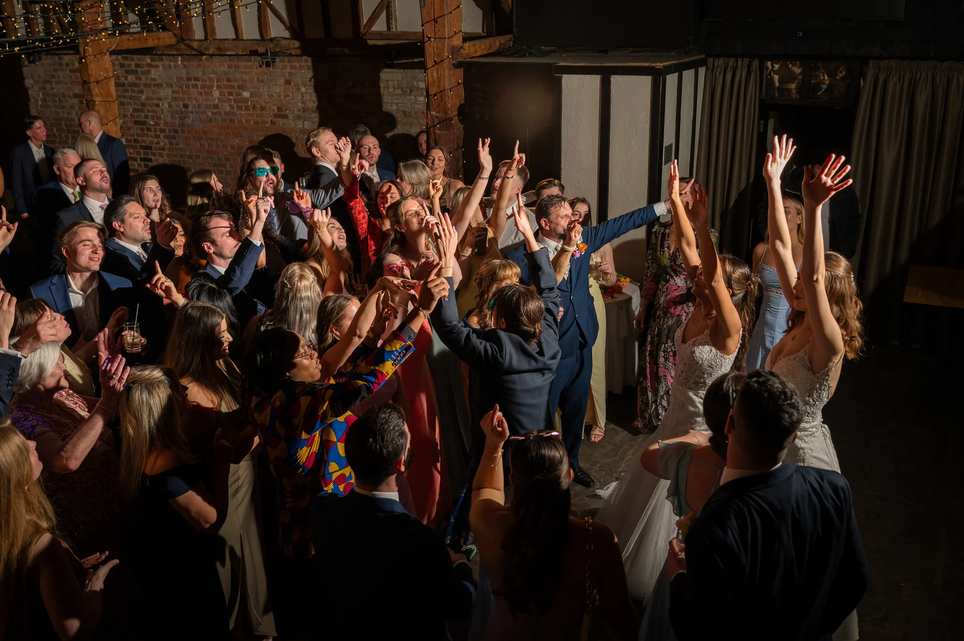 An energetic crowd of wedding guests dances enthusiastically on a packed dance floor under a rustic barn ceiling with exposed brick and twinkling fairy lights.