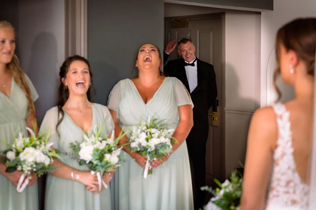 Bridesmaids in sage dresses laugh joyfully as they and the groom share a first look with the bride in her wedding dress.