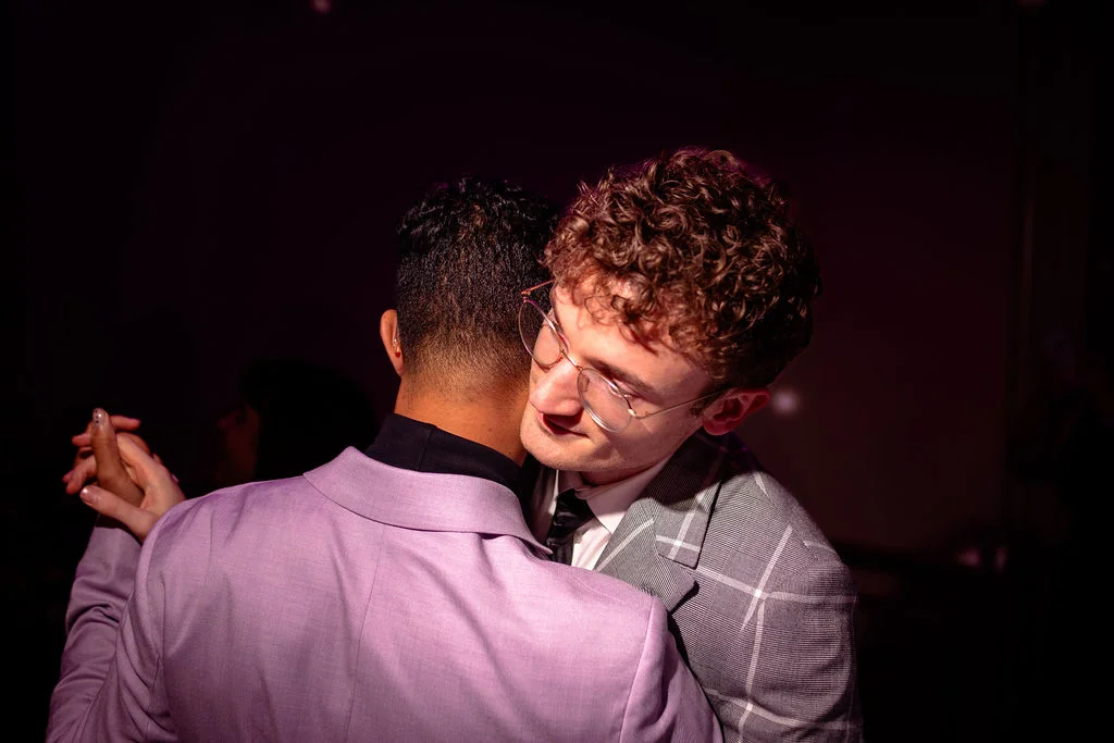 Two grooms share a tender embrace while dancing together during their evening reception, with one groom wearing a lilac suit and the other in a grey checked jacket.