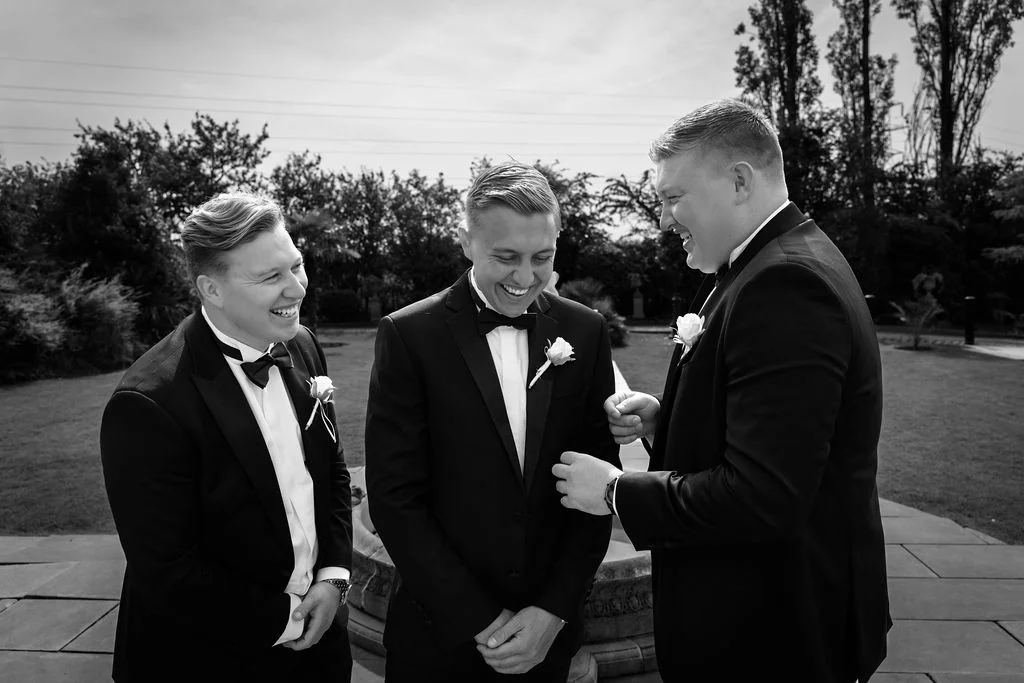 Three groomsmen in black tuxedos share a joyful laugh together outdoors while standing on a paved patio.