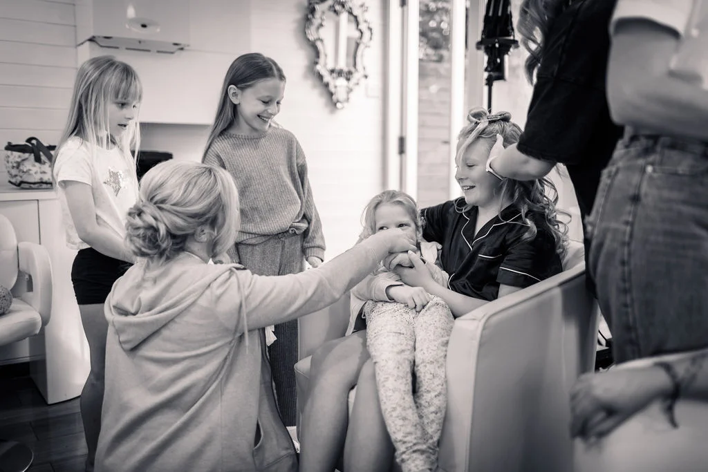 A candid black and white shot captures a group of children and women laughing together during the morning hair styling preparations.