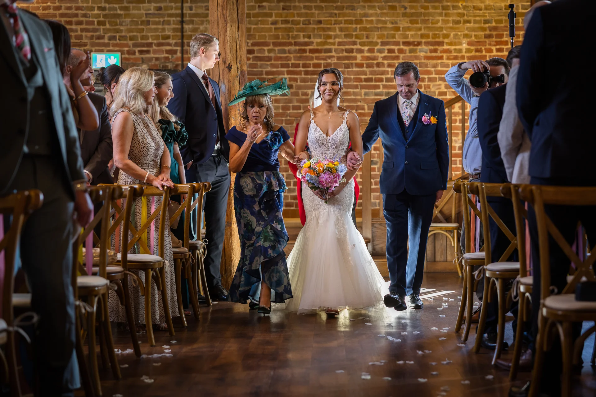 A smiling bride walks down the aisle in a rustic brick barn venue, escorted by her father and mother while holding a colourful bouquet.