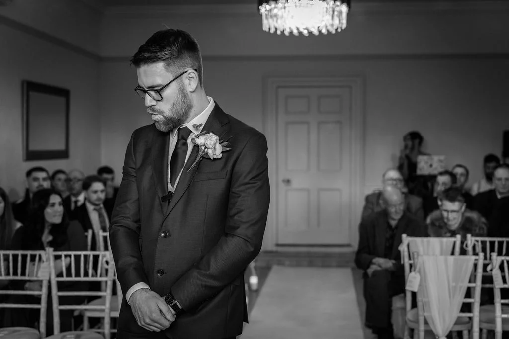 An emotional groom stands at the altar waiting for his bride during an indoor civil ceremony, wearing a tailored dark suit and buttonhole.