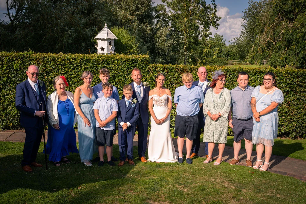 A large wedding party and family group pose together outdoors in a sunlit garden with a neat green hedge backdrop.