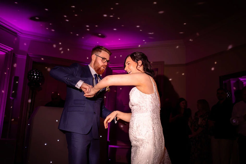 A newlywed couple laugh and dance joyfully together under vibrant purple lighting and scattered reflection dots during their evening reception.
