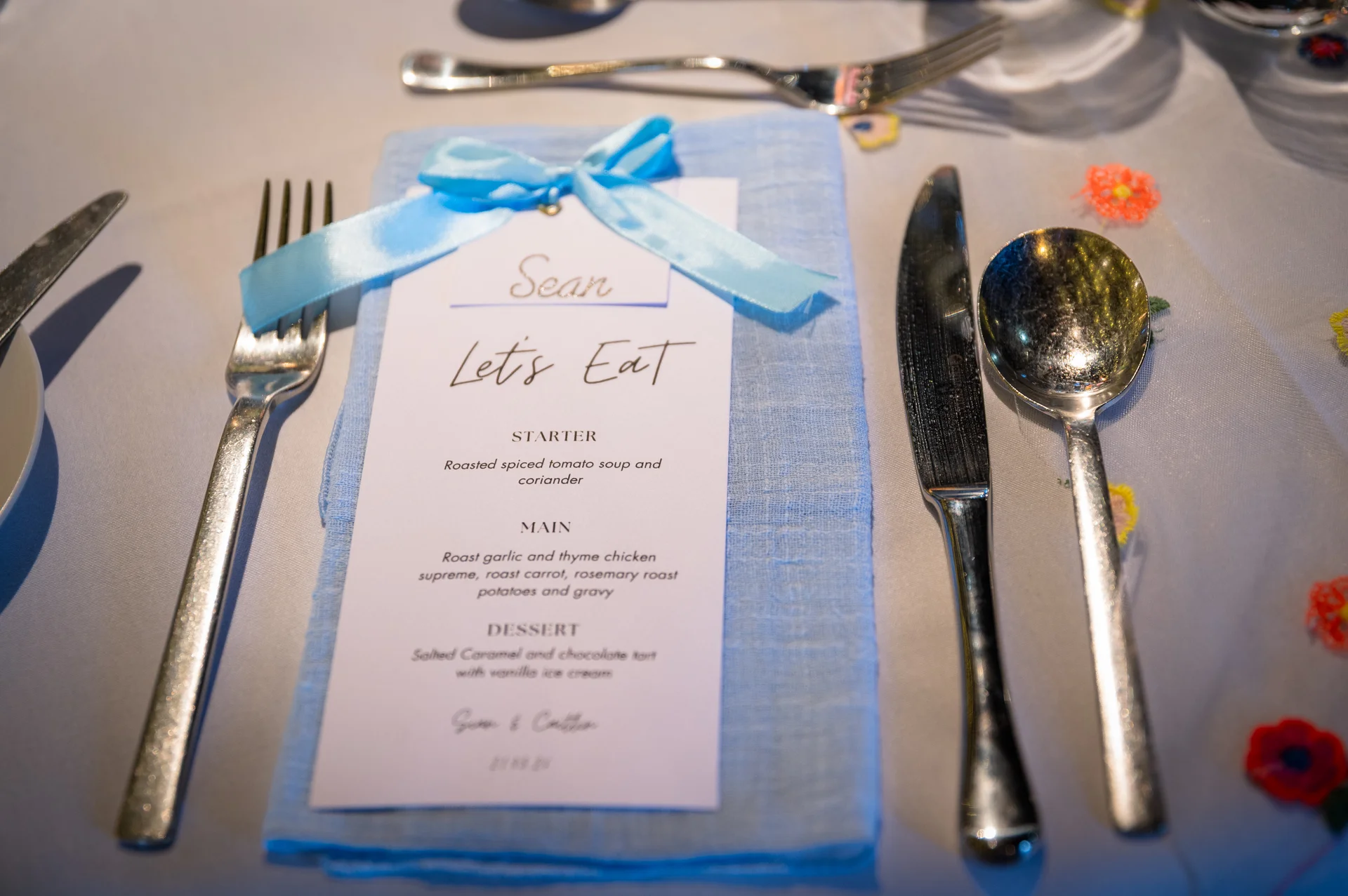 A close up of a wedding place setting featuring a light blue linen napkin topped with a personalised menu card tied with a pale blue ribbon.